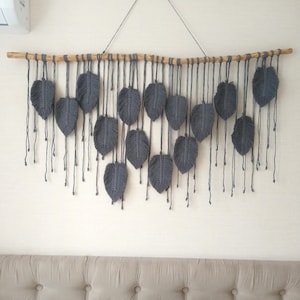 May include: Gray macrame wall hanging with a wooden dowel and a series of gray feather shapes hanging from it.