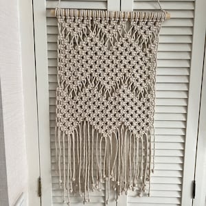 Puede incluir: Un tapiz de macramé beige con un patrón geométrico y flecos. El tapiz está hecho de cuerda de fibra natural y está sujeto a una varilla de madera.