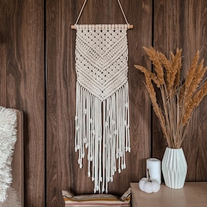 Op de afbeelding: Een crèmekleurige macramé wandhanger met een geometrisch patroon en franjes. De wanddecoratie hangt aan een houten stok tegen een donkere houten wand. Decoratieve elementen zijn een witte vaas met gedroogde planten en een witte pompoen.