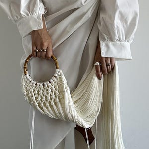 Pode incluir: Uma bolsa de mão de macramê branco com alça de bambu e longas franjas. A bolsa é segurada por uma pessoa vestindo um vestido branco de mangas compridas. As franjas da bolsa pendem, criando um efeito fluido.