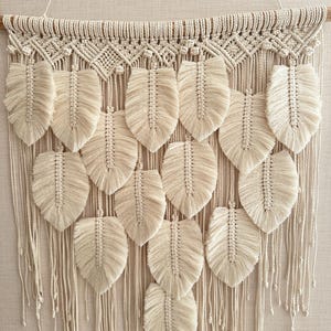 May include: Cream-colored macrame wall hanging featuring a wooden dowel, intricate knotted top, and multiple feather-shaped elements. The feathers are arranged in rows, with long, dangling fringe below. A decorative, bohemian-style piece.