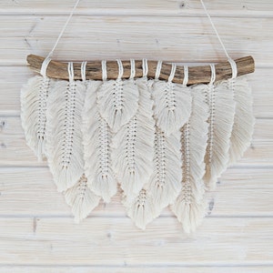 Puede incluir: Un tapiz de macramé de pared con una rama de madera natural y cordón de algodón blanco, creando un diseño de plumas.
