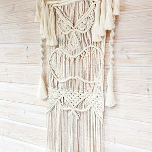 Puede incluir: Tapiz de macramé de color crema con intrincados patrones geométricos y borlas. El textil hecho a mano presenta una serie de diseños anudados, incluyendo formas de diamante y corazón, y está suspendido de una sección superior tejida. La decoración de pared se muestra sobre un fondo de madera clara.