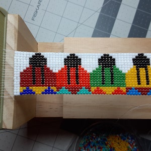 Könnte beinhalten: Ein Perlenarmband, das auf einem Webstuhl hergestellt wird. Das Armband zeigt ein Muster aus vier bunten, geometrischen Formen mit schwarzen Umrissen. Die Formen sind rot, orange, grün und gelb.