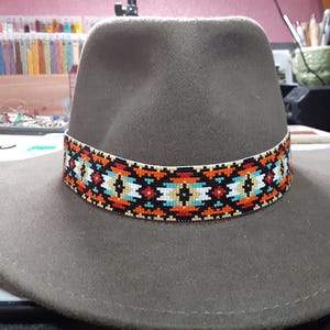 May include: A grey fedora hat with a decorative band featuring a geometric pattern in orange, black, white, and turquoise. The hat has a wide brim and a classic design.