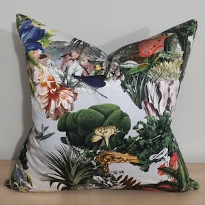 Op de afbeelding: Een wit kussen met een bloemen- en bladprint. De print bevat een verscheidenheid aan bloemen, bladeren en takken in tinten groen, rood, roze en blauw.