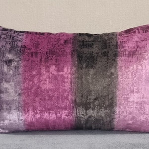Puede incluir: Una almohada rectangular con un patrón de rayas en tonos de morado, gris y negro. La almohada tiene una textura suave y aterciopelada.
