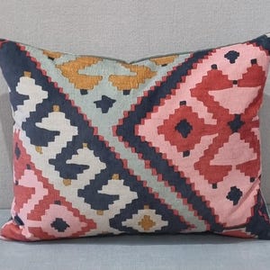 May include: Rectangular decorative pillow with a geometric pattern in shades of navy, pink, red, gold, and white. The pillow has a soft, textured appearance and is likely made of fabric. The design features repeating diamond and zigzag shapes.