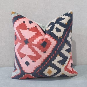May include: Square decorative pillow with a geometric pattern in shades of pink, red, navy, and cream. The pillow has a light blue backing and a soft, textured appearance. The design features a repeating diamond and zigzag motif.