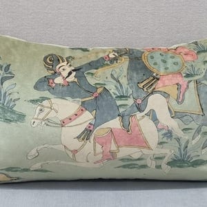 May include: Rectangular decorative pillow with a light green background and a detailed illustration of figures on horseback. The figures are dressed in blue, pink, and white, with a landscape in the background.