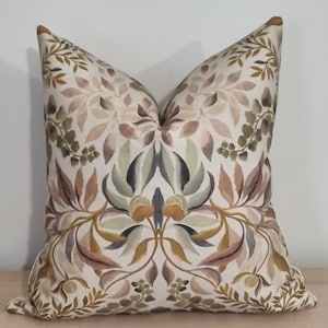 Puede incluir: Una almohada decorativa con un patrón floral en tonos marrones, verdes y rosas sobre un fondo crema.