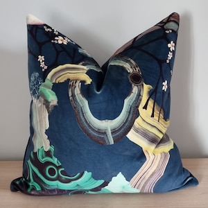 May include: A square decorative pillow with a dark blue background and a unique abstract design. The design features a mix of colors including green, yellow, and brown, with white floral accents. The pillow is made of a soft, velvety material.