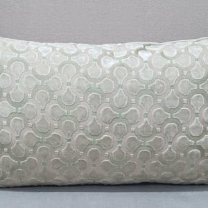 May include: A rectangular decorative pillow with a repeating geometric pattern in shades of light gray and mint green. The pillow has a soft, textured appearance, and is placed on a gray surface.