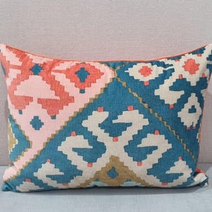 May include: Rectangular decorative pillow with a geometric pattern in shades of pink, teal, white, and coral. The pillow has a coral-colored trim and a soft, textured appearance, suitable for home decor.