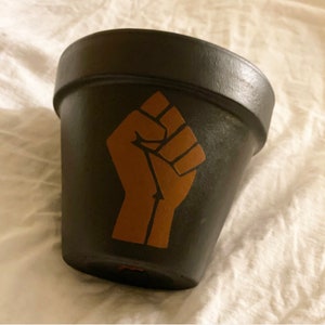 May include: Black terracotta flower pot with a brown raised fist design.