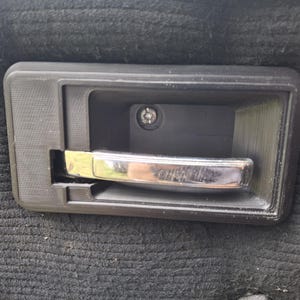 Może przedstawiać: Czarny uchwyt do drzwi samochodu z chromowanym uchwytem. Uchwyt jest osadzony w czarnej plastikowej ramie, ze śrubą widoczną we wnęce. Uchwyt jest zamontowany na czarnej, teksturowanej powierzchni.