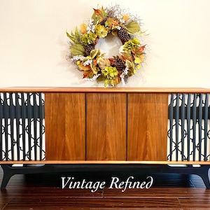 Peut inclure: Une console stéréo vintage de style moderne du milieu du siècle avec des grilles de haut-parleurs à motifs géométriques noirs et blancs, des portes en placage de bois et des pieds noirs. La console est posée sur un sol en bois. Le texte "Vintage Refined" est visible en bas au centre de l'image.