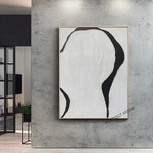 May include: Abstract art piece featuring a black, organic shape against a white background. The artwork is framed in a gold-colored border and mounted on a gray wall. The painting has a textured appearance, with visible brushstrokes. The artwork is minimalist in style, with a focus on form and contrast.