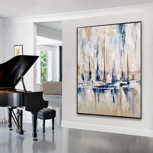 May include: Abstract art painting with a black frame. The artwork features vertical brushstrokes in shades of blue, beige, and white, creating a textured effect. The painting is displayed in a bright, modern interior setting.