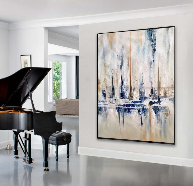 May include: Abstract art painting with a black frame. The artwork features vertical brushstrokes in shades of blue, beige, and white, creating a textured effect. The painting is displayed in a bright, modern interior setting.