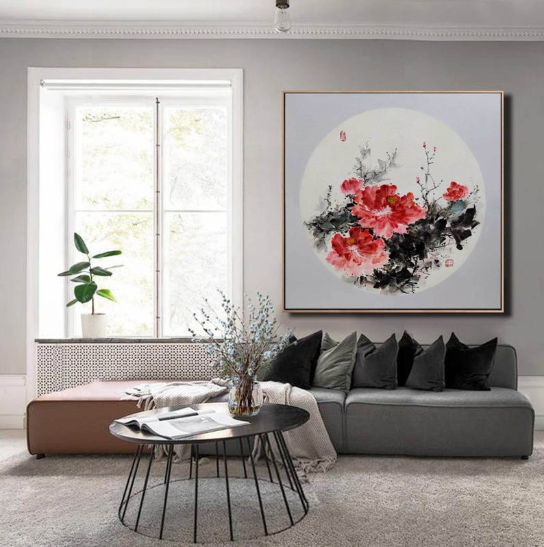 Puede incluir: Pintura enmarcada que representa peon&iacute;as rojas con hojas negras y verdes, sobre un fondo circular. La obra se exhibe en un sal&oacute;n con un sof&aacute; gris y una mesa de centro oscura. El marco es de color dorado.