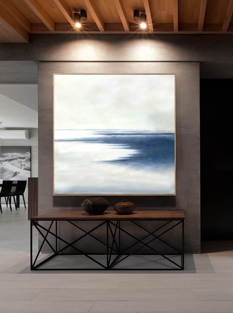 May include: An abstract painting with a gold frame depicts a seascape in shades of blue and white. The artwork hangs above a wooden console table with a black metal base. Two decorative ceramic vases sit on the table.