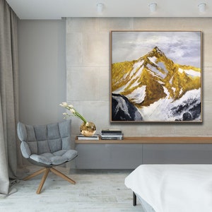 May include: A large abstract painting of a mountain range with gold and white accents. The painting is framed in a thin, gold frame and is hanging on a wall in a modern living room.