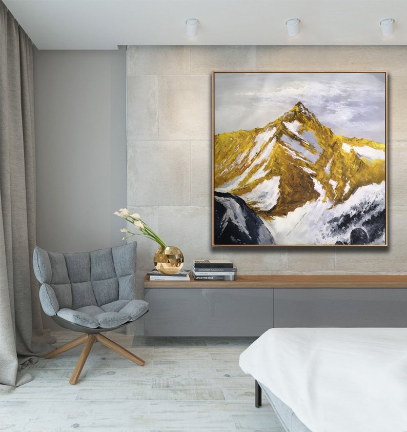 May include: A large abstract painting of a mountain range with gold and white accents. The painting is framed in a thin, gold frame and is hanging on a wall in a modern living room.