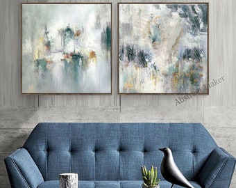 Könnte beinhalten: Zwei abstrakte Gemälde in gedeckten Farben von Grün, Grau und Weiß. Die Gemälde sind gerahmt und an einer grauen Wand über einem blauen Sofa aufgehängt.