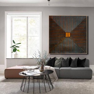 May include: A square abstract painting with a geometric design of intersecting lines in shades of brown, gray, and orange. The painting is hanging on a wall in a living room setting.