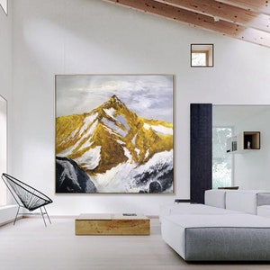 May include: Abstract painting of a mountain range with gold and white accents. The painting is framed and hanging on a white wall in a modern living room.