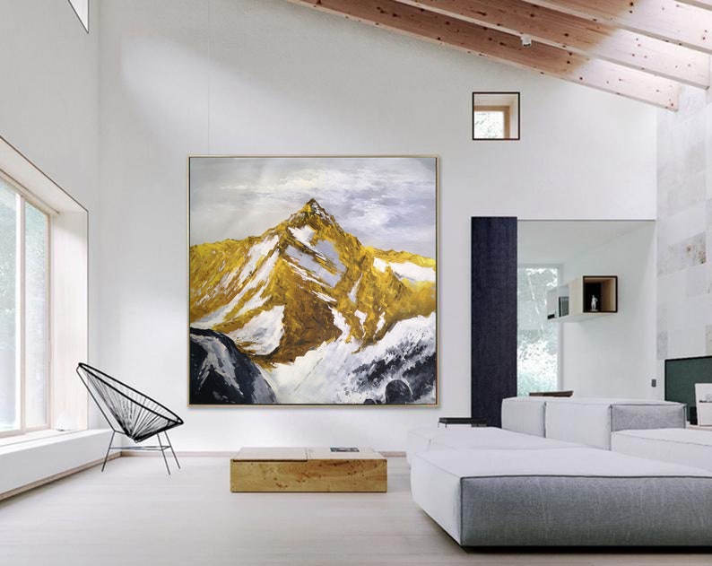 May include: Abstract painting of a mountain range with gold and white accents. The painting is framed and hanging on a white wall in a modern living room.