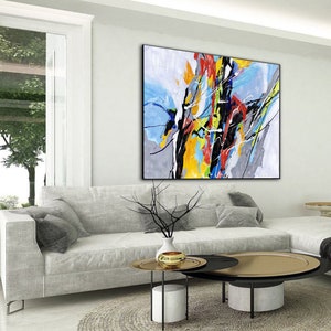 May include: Abstract painting with bold strokes of yellow, blue, red, and black on a gray background. The painting is hanging on a white wall in a living room setting.