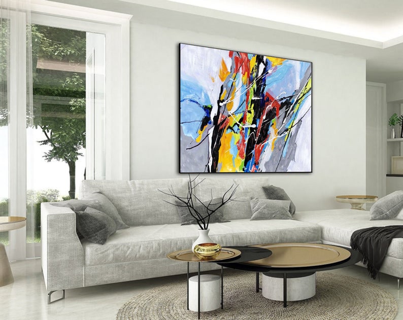 May include: Abstract painting with bold strokes of yellow, blue, red, and black on a gray background. The painting is hanging on a white wall in a living room setting.