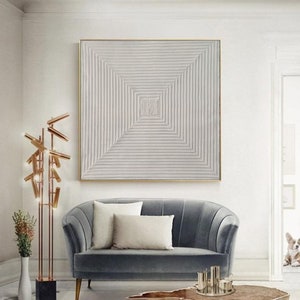 May include: A white abstract painting with a geometric pattern of lines creating a square in the center. The painting is framed in a thin gold frame and hangs on a white wall above a gray velvet sofa.