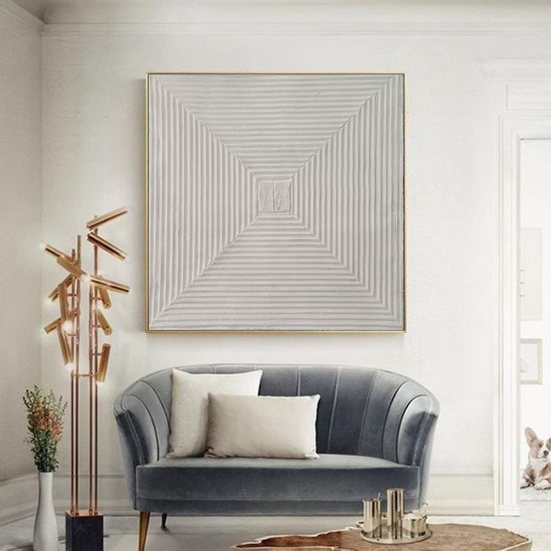 May include: A white abstract painting with a geometric pattern of lines creating a square in the center. The painting is framed in a thin gold frame and hangs on a white wall above a gray velvet sofa.