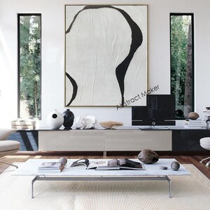 May include: Large abstract painting with a white textured background and bold black curved lines. The artwork is displayed in a modern living room setting, above a low cabinet. The room features minimalist furniture and decor.