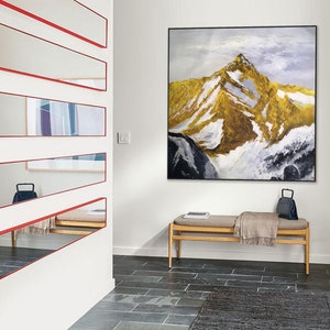 May include: A hallway with a white wall and a long, narrow mirror with a red frame. The mirror is divided into five sections. There is a painting of a mountain range on the wall opposite the mirror. A wooden bench with a blanket and a black bag sits in front of the painting.