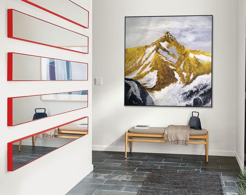 May include: A hallway with a white wall and a long, narrow mirror with a red frame. The mirror is divided into five sections. There is a painting of a mountain range on the wall opposite the mirror. A wooden bench with a blanket and a black bag sits in front of the painting.
