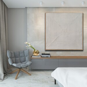 May include: A white textured square canvas painting with a geometric pattern of raised lines in a square shape. The painting is framed in a thin, light wood frame and is hanging on a wall in a modern living room.
