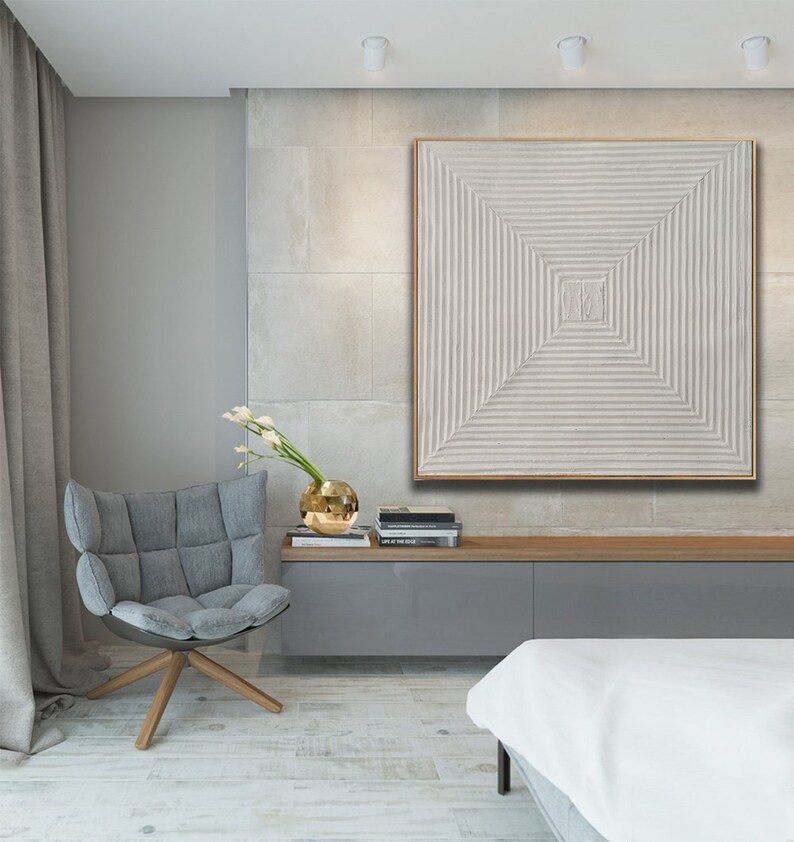 May include: A white textured square canvas painting with a geometric pattern of raised lines in a square shape. The painting is framed in a thin, light wood frame and is hanging on a wall in a modern living room.