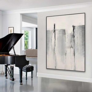 May include: Abstract painting with gray and white brushstrokes on a white canvas. The painting is framed in a black frame and is hanging on a white wall in a modern living room.