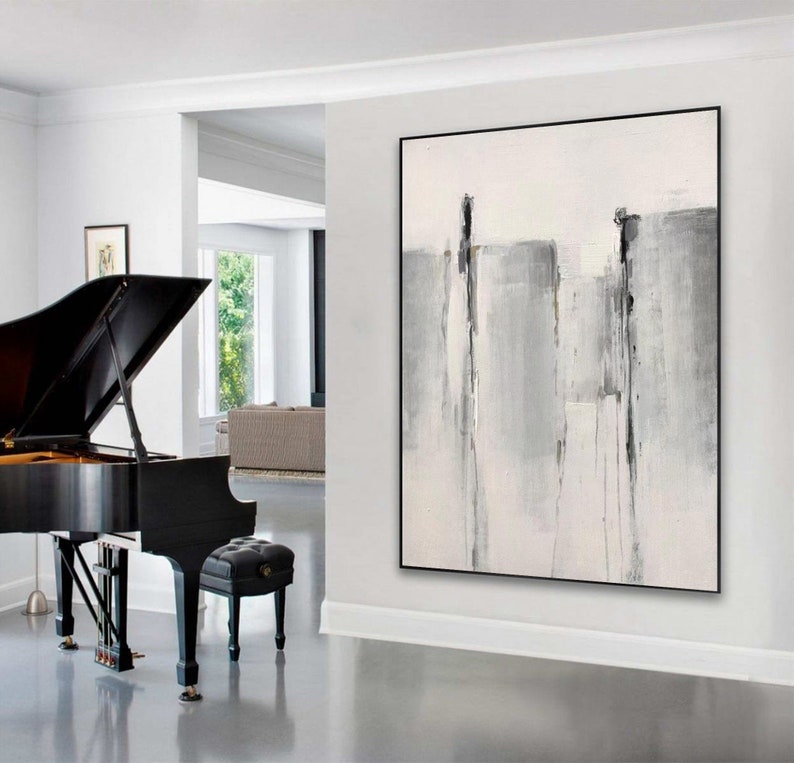 May include: Abstract painting with gray and white brushstrokes on a white canvas. The painting is framed in a black frame and is hanging on a white wall in a modern living room.