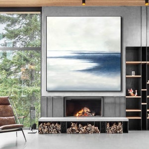 May include: Large abstract painting with a minimalist design. The artwork features a serene landscape with a gradient of white and blue hues, framed in a black border. The painting hangs above a fireplace, enhancing the room's modern aesthetic.