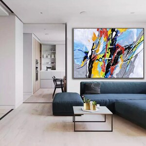 May include: Abstract painting with bold strokes of black, yellow, blue, red, and white paint on a gray and white background. The painting is framed in a thin gold frame and is hanging on a wall in a living room.