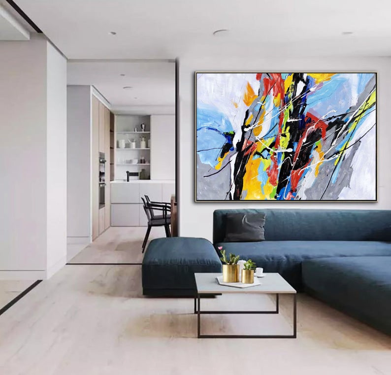 May include: Abstract painting with bold strokes of black, yellow, blue, red, and white paint on a gray and white background. The painting is framed in a thin gold frame and is hanging on a wall in a living room.