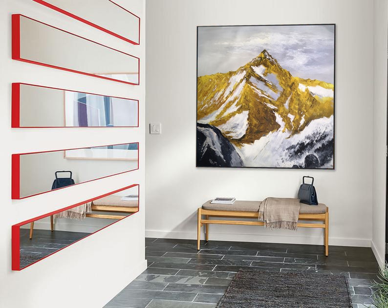 May include: A hallway with a white wall and a wooden bench. The wall has five horizontal mirrors with red frames. A large framed painting of a mountain range with snow and gold accents hangs on the wall.