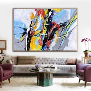 May include: Abstract painting with bold strokes of yellow, blue, red, black, and gray on a white background. The painting is framed and hanging on a white wall in a living room setting.