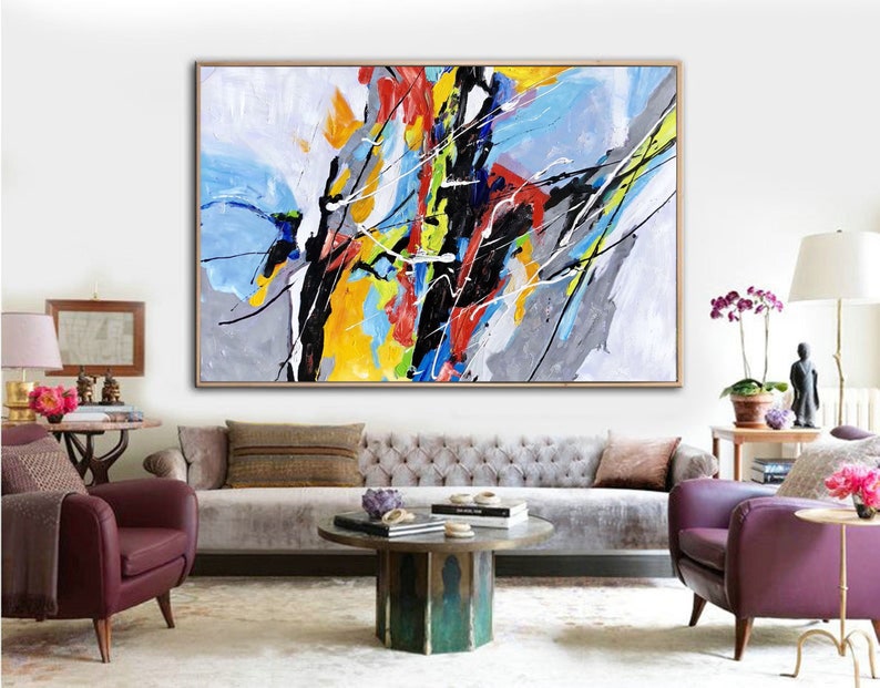 May include: Abstract painting with bold strokes of yellow, blue, red, black, and gray on a white background. The painting is framed and hanging on a white wall in a living room setting.