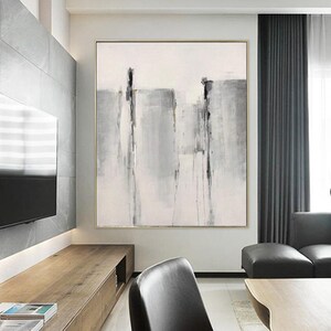 May include: Abstract painting with gray and white brushstrokes on a white canvas. The painting is framed in a thin gold frame and is hanging on a wall in a modern living room.